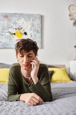 Sıradan giyinmiş üzgün kıvırcık travesti yatak odasında akıllı telefondan konuşuyor, telefon görüşmesi yapıyor.
