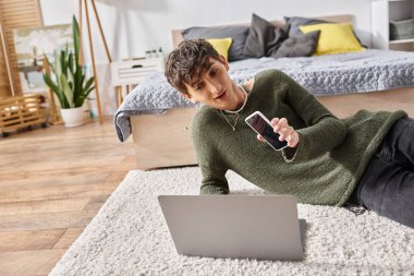 Mutlu transseksüel etkileyicisi dizüstü bilgisayar kullanıyor ve halının üzerinde otururken akıllı telefon tutuyor, blog yazarı