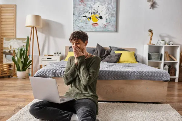 stock image shocked and curly transgender influencer covering mouth and using laptop and sitting near bed
