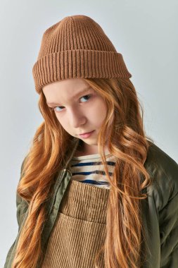 serious preteen girl in knitted hat and winter outerwear looking at camera on grey backdrop clipart