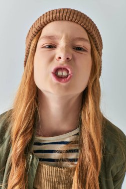angry preteen girl in knitted hat and outerwear showing teeth and looking at camera on grey backdrop clipart