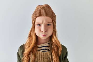 displeased preteen girl in stylish winter outfit with knitted hat puffing cheeks on grey background clipart