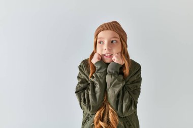 preteen girl in stylish winter outfit with knitted hat feeling cold while standing on grey backdrop clipart