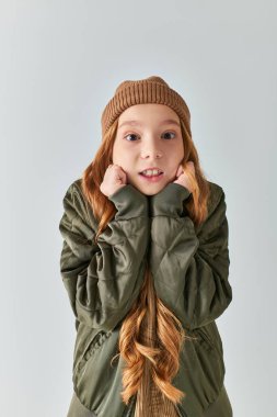 girl in winter outfit with knitted hat feeling cold and standing on grey backdrop, looking at camera clipart