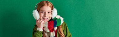 preteen girl in ear muffs, striped scarf and winter outfit smiling on turquoise backdrop, banner clipart