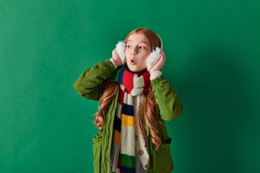 shocked preteen girl in ear muffs, striped scarf and winter outfit looking away on turquoise clipart