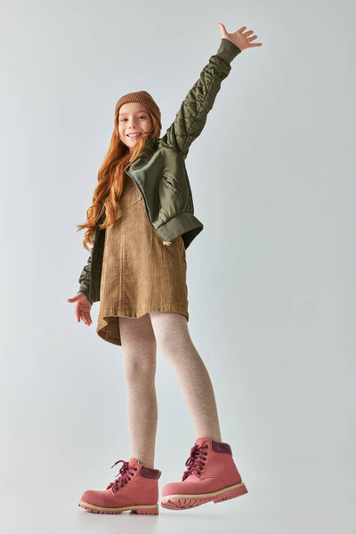 веселая девушка в стильном зимнем наряде в сапогах и шляпе улыбается и машет рукой на сером фоне