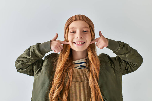 positive preteen girl in knitted hat and winter outfit pointing at cheeks with fingers on grey