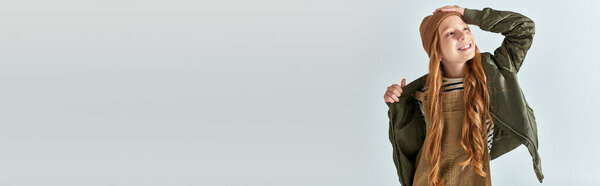 довольная девочка-подросток в трикотажной шляпе и теплая одежда, регулирующая зимнюю куртку на сером, баннер
