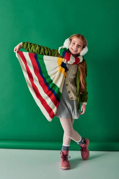 stock image positive kid in ear muffs and scarf standing in winter outfit on turquoise backdrop, cozy layers