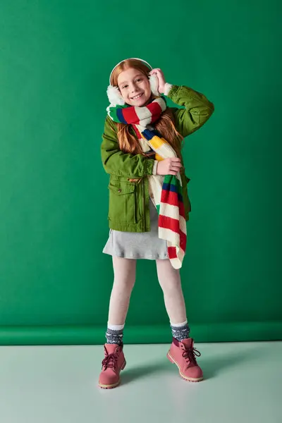 stock image positive child in ear muffs and scarf standing in winter outfit on turquoise backdrop, cozy layers