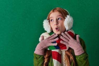 Şok olmuş kulaklıklı genç kız, çizgili atkı ve turkuaz arka planda el kol hareketi yapan kışlık kıyafet.