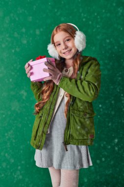 preteen girl in ear muffs, scarf and winter attire holding Christmas present under falling snow clipart