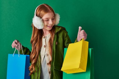holiday season concept, happy kid in winter outfit and ear muffs looking at shopping bags with gifts clipart