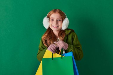 holiday season, excited kid in winter outfit and ear muffs holding shopping bags on turquoise clipart