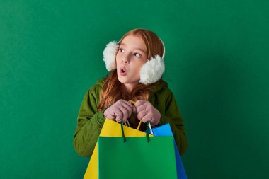 winter holidays, excited kid in winter outfit and ear muffs holding shopping bags on turquoise clipart