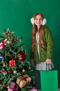 winter holidays, joyful girl in ear muffs holding shopping bag near Christmas tree, falling snow clipart