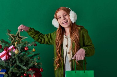 winter holidays, excited girl in ear muffs holding shopping bag touching star top of Christmas tree clipart