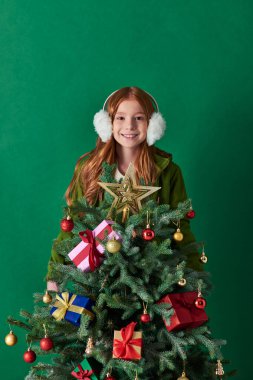 winter holidays, happy girl in ear muffs standing behind decorated Christmas tree on turquoise clipart