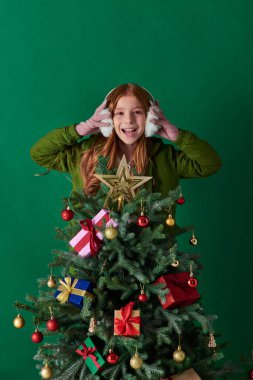 holidays, excited girl wearing ear muffs and standing behind decorated Christmas tree on turquoise clipart