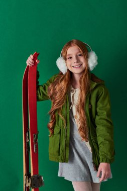 happy preteen girl in ear muffs and winter outfit smiling and holding red skis on turquoise backdrop clipart