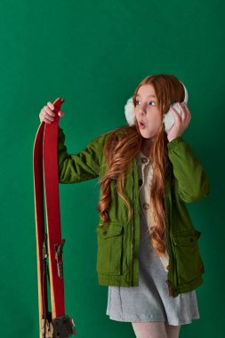 amazed preteen girl in ear muffs and winter outfit holding red ski gear on turquoise backdrop clipart