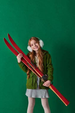 cheerful preteen girl in ear muffs and winter outfit holding red ski gear on turquoise backdrop clipart