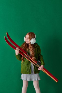 joyful preteen girl in ear muffs and winter outfit holding red ski gear on turquoise backdrop clipart