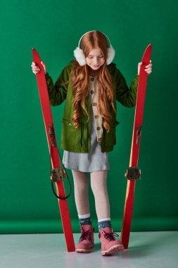 joyful preteen girl in ear muffs and winter attire holding red skis on turquoise background clipart