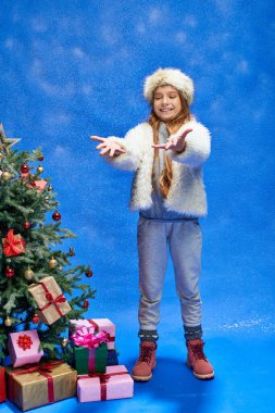 Sahte kürk ceketli ve şapkalı mutlu kız Noel ağacının yanında duruyor mavi, kar üzerinde hediyelerle.