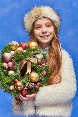 Sahte kürk şapkalı ve ceketli mutlu kız mavi zeminde kar altında Noel çelengi tutuyor.