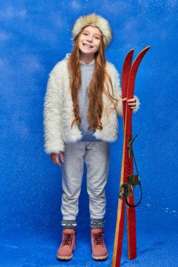 cheerful preteen girl in winter faux fur jacket and hat holding red skis on turquoise background clipart