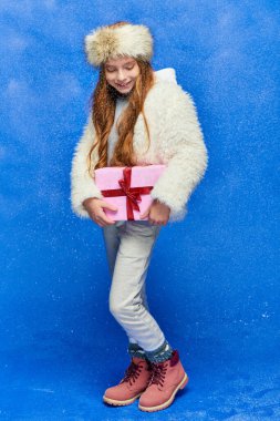 winter joy, pleased preteen girl in faux fur jacket and hat holding gift box on turquoise background clipart