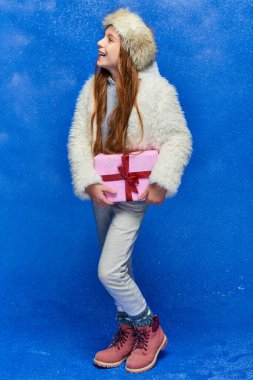 winter joy, happy preteen girl in faux fur jacket and hat holding gift box on turquoise background clipart