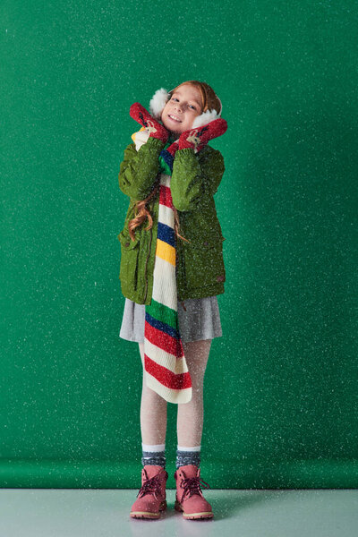 счастливая девушка в наушниках, теплый шарф и зимний наряд стоя под падающим снегом на бирюзовой