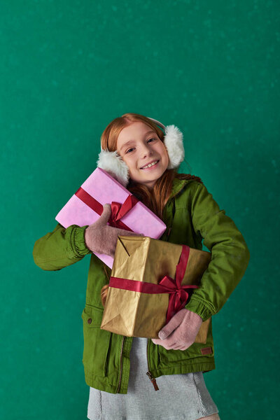 holiday season, happy kid in winter outfit and ear muffs holding holiday gifts under falling snow