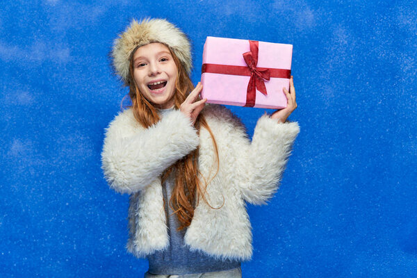 winter holidays, cheerful girl in faux fur jacket and hat holding gift box on turquoise background