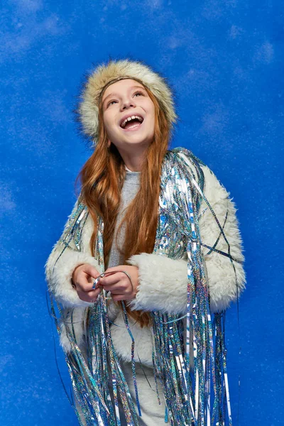 stock image excited girl in faux fur jacket and hat with tinsel standing under falling snow on blue backdrop