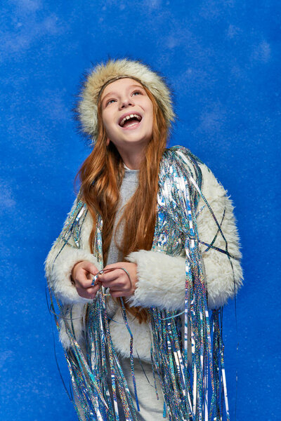 excited girl in faux fur jacket and hat with tinsel standing under falling snow on blue backdrop