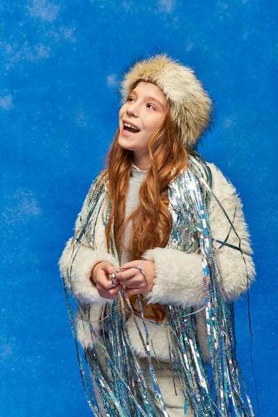 cheerful girl in faux fur jacket and hat with tinsel standing under falling snow on blue backdrop