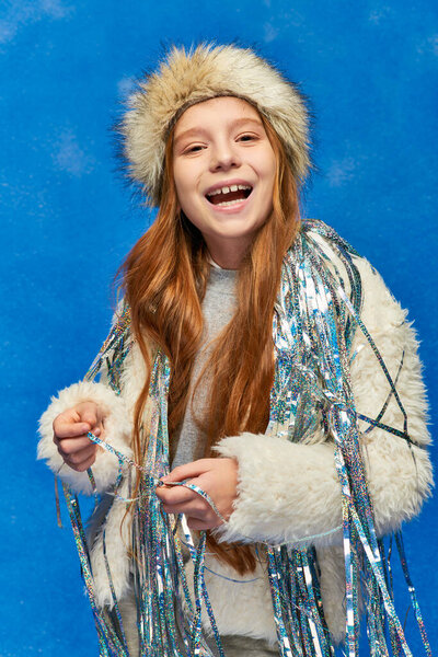 smiling girl in faux fur jacket and hat with tinsel standing under falling snow on blue backdrop
