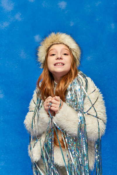 girl in faux fur jacket with tinsel standing under falling snow on blue backdrop, feeling cold
