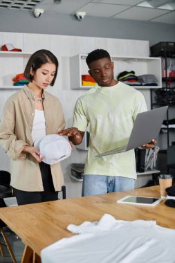 Dizüstü bilgisayarı ve beyaz şapkası olan çok ırklı moda tasarımcıları baskı stüdyosunda yeni projeyi tartışıyor