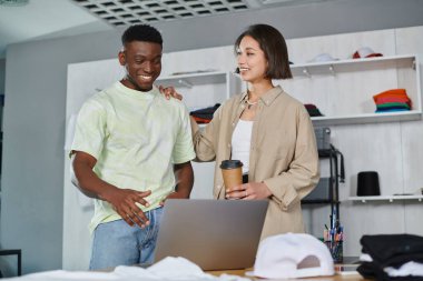 pleased asian woman touching shoulder of african american colleague near laptop in print studio clipart