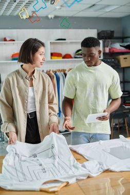 young interracial small business owners talking near t-shirts with format sizes in print studio clipart