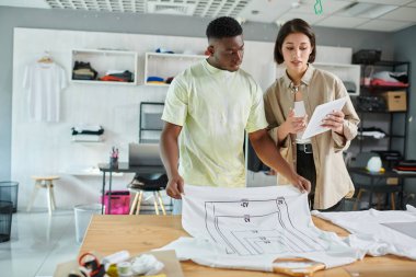 Irklar arası tasarımcılar baskı stüdyosunda format boyutunda tişörtlerin yakınındaki dijital tablete bakıyorlar.