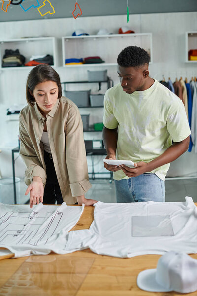asian woman pointing at t-shirt with format sizes near african american designer with digital tablet