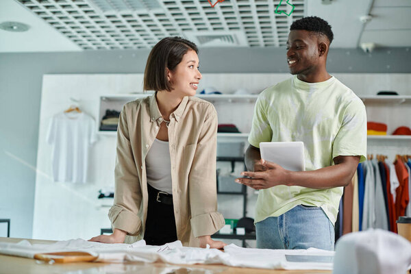 african american designer with digital tablet talking to asian woman near clothes in print studio