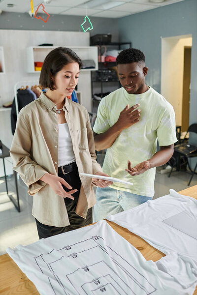african american designer looking at digital tablet and talking to asian colleague in print studio