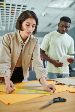 asian fashion designer measuring sewing pattern near african american colleague in print studio clipart